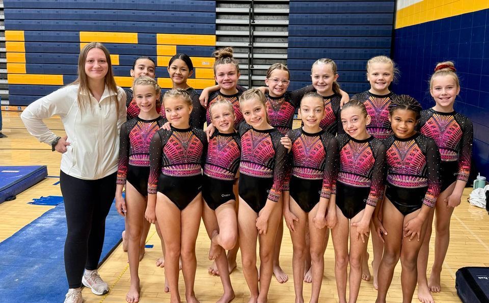 Gym team posing in matching black-and-pink uniforms with their coach in a gymnasium.
