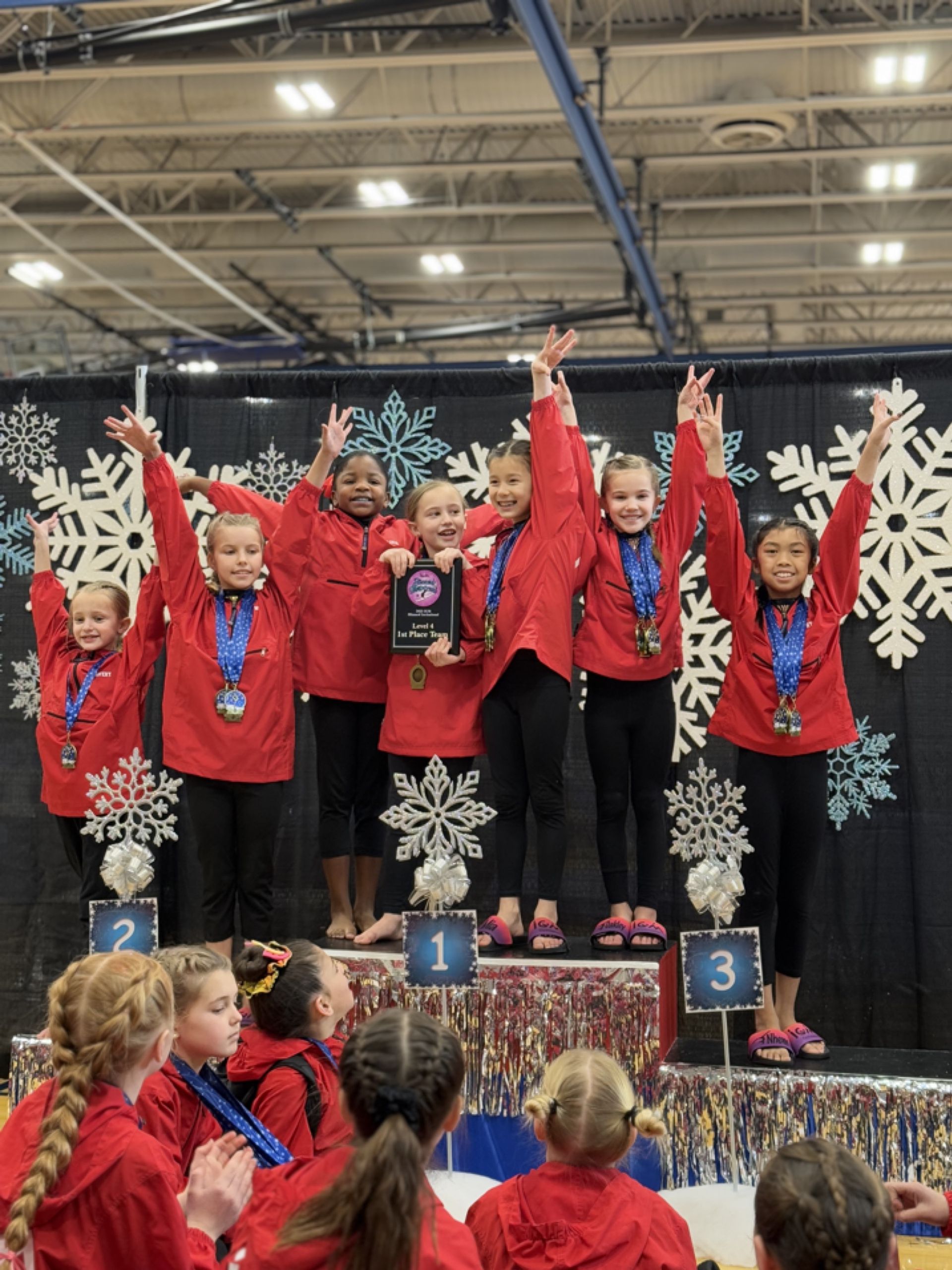 Gymnastics team in red jackets on a podium, raising medals and a trophy at an indoor award ceremony