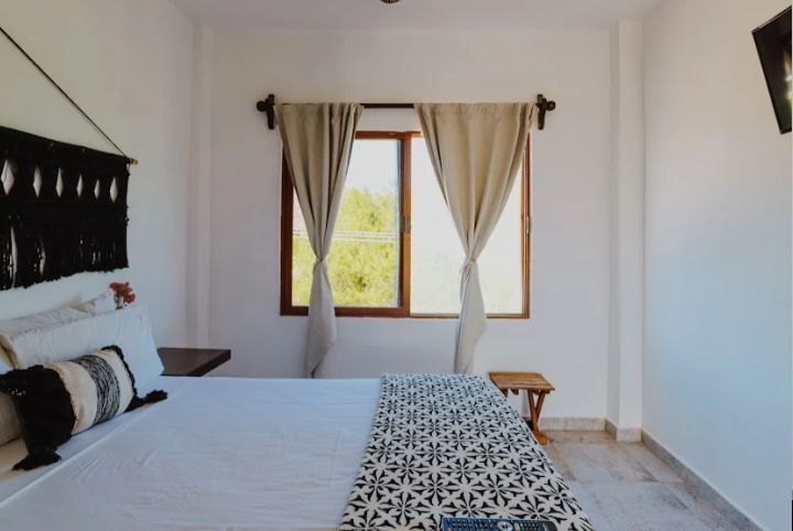 Habitación luminosa con cama, manta estampada, ventana con cortinas y paredes blancas.