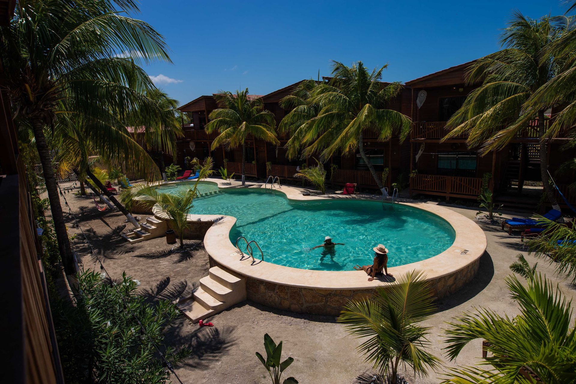 Piscina de un complejo tropical con dos nadadores, palmeras y tumbonas bajo un sol radiante.