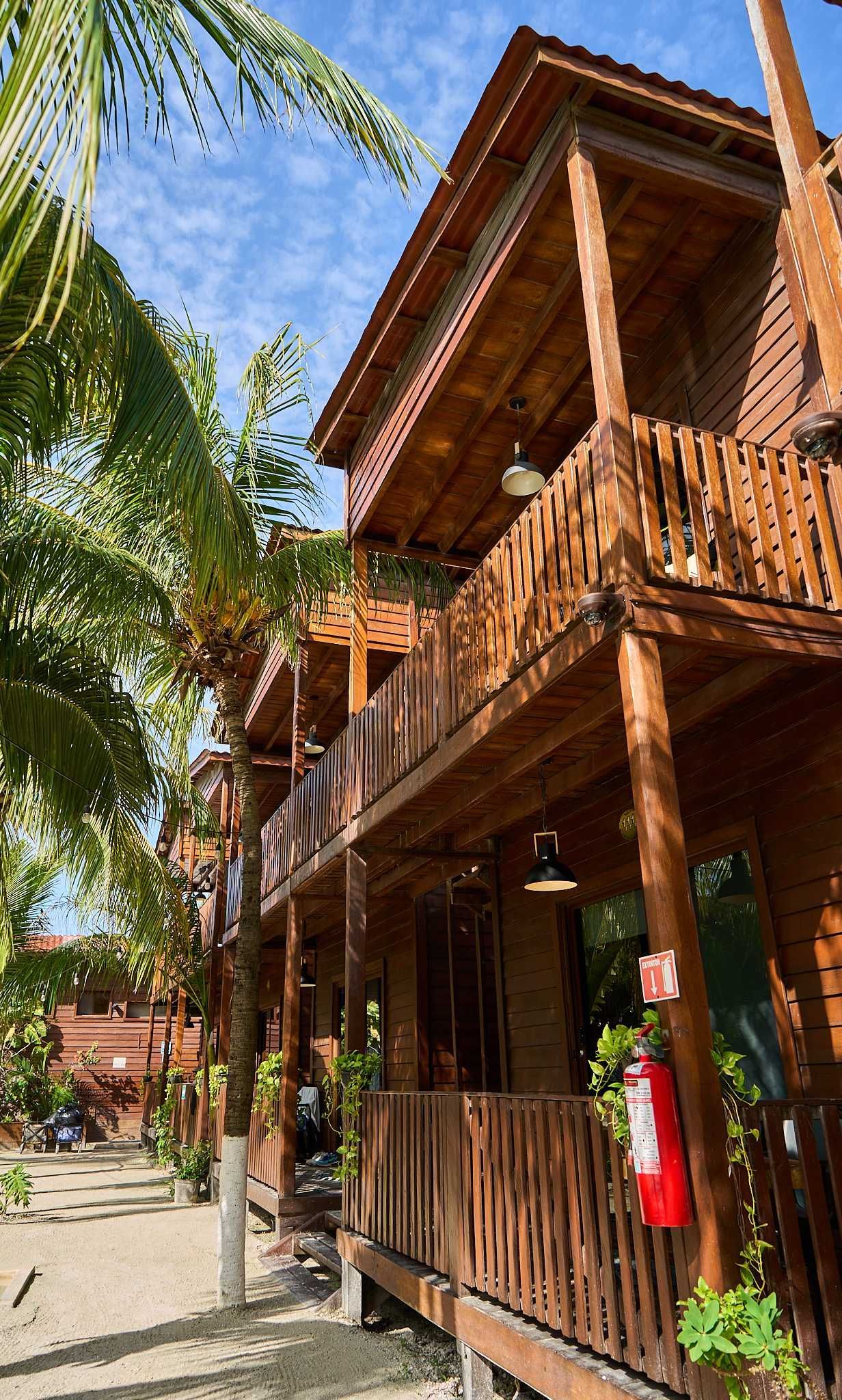 Cabaña tropical de madera con balcones y palmeras a lo largo de un sendero soleado.