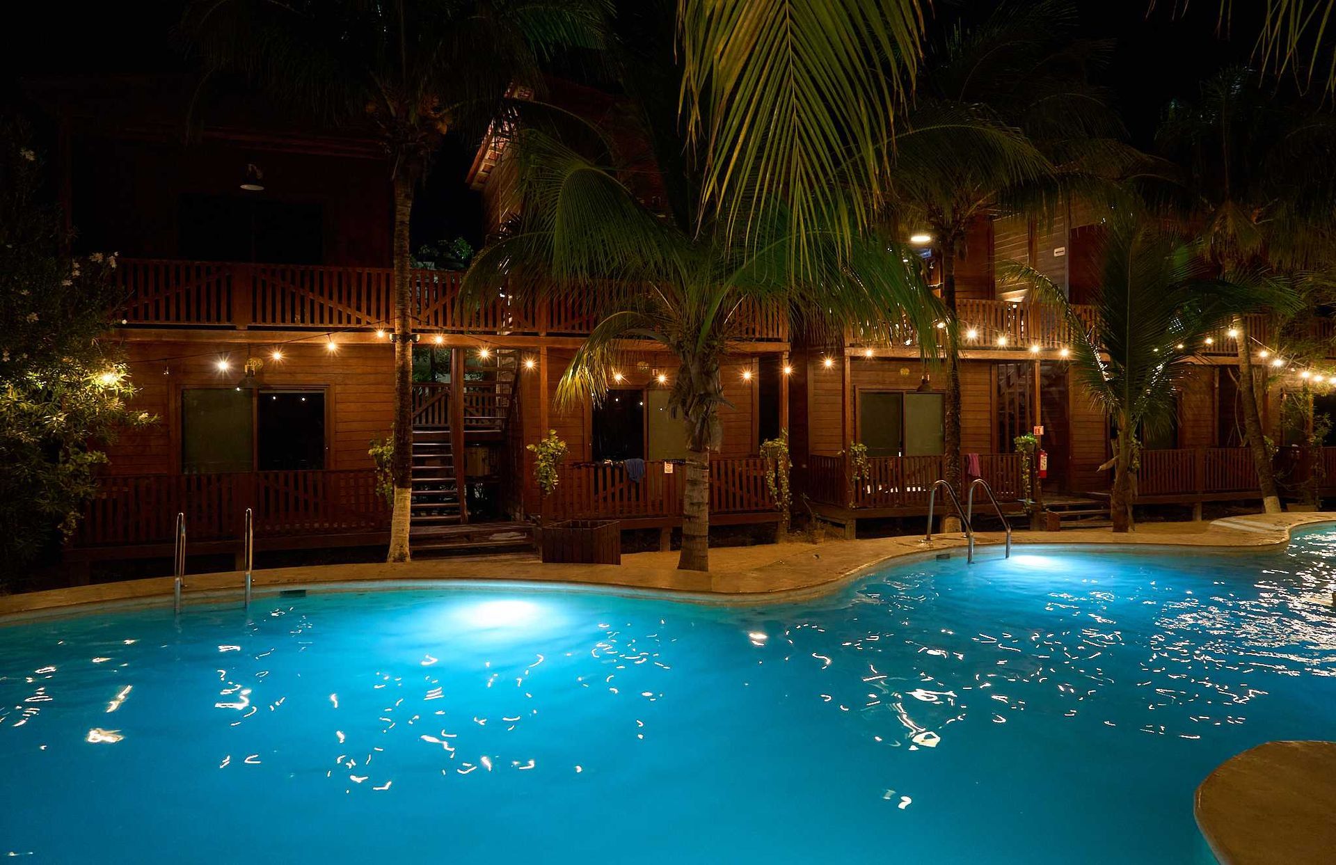 Piscina iluminada con luz azul por la noche, junto a un patio del complejo turístico bordeado de palmeras.