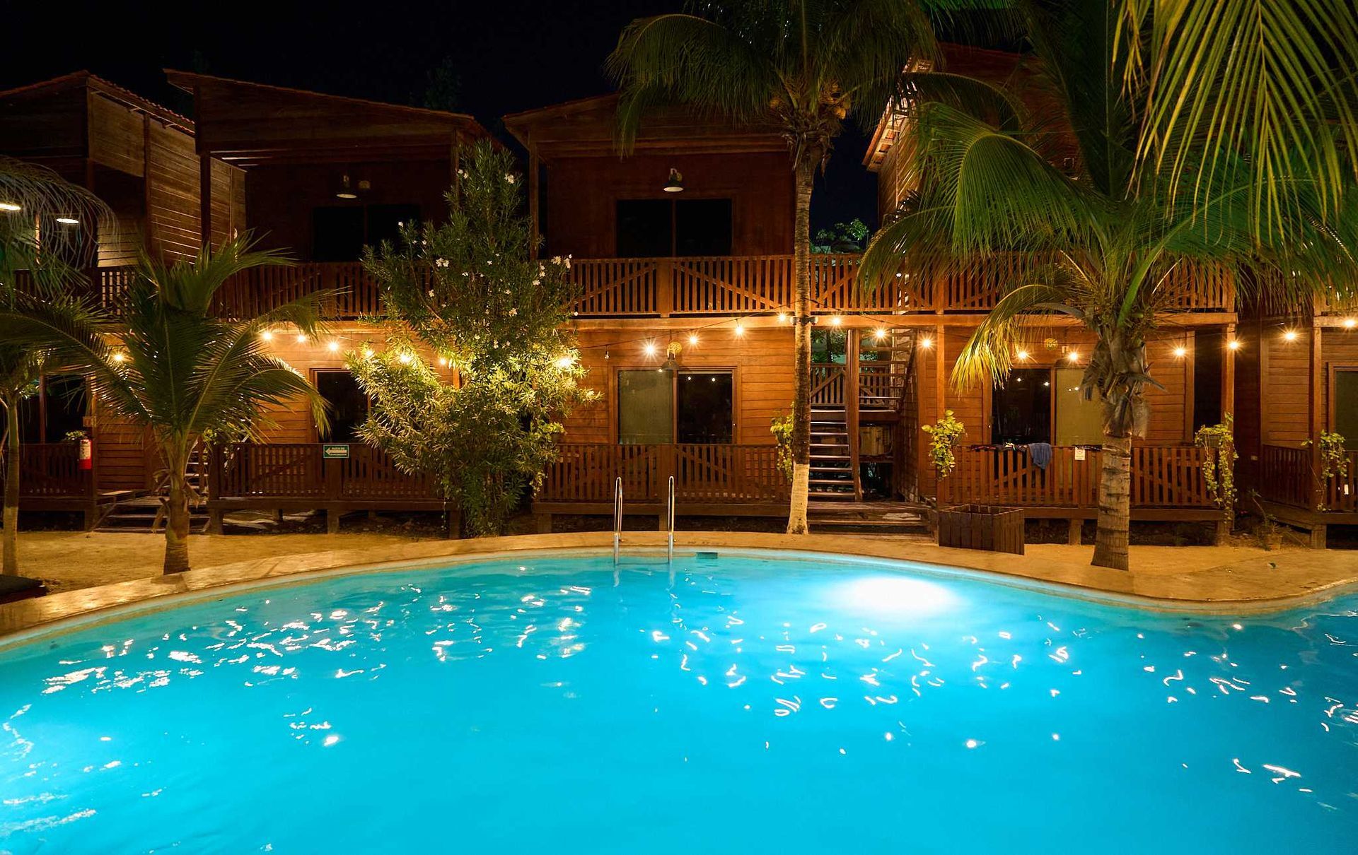 Piscina nocturna de un complejo turístico con agua azul brillante, palmeras y edificios cálidamente iluminados al fondo.