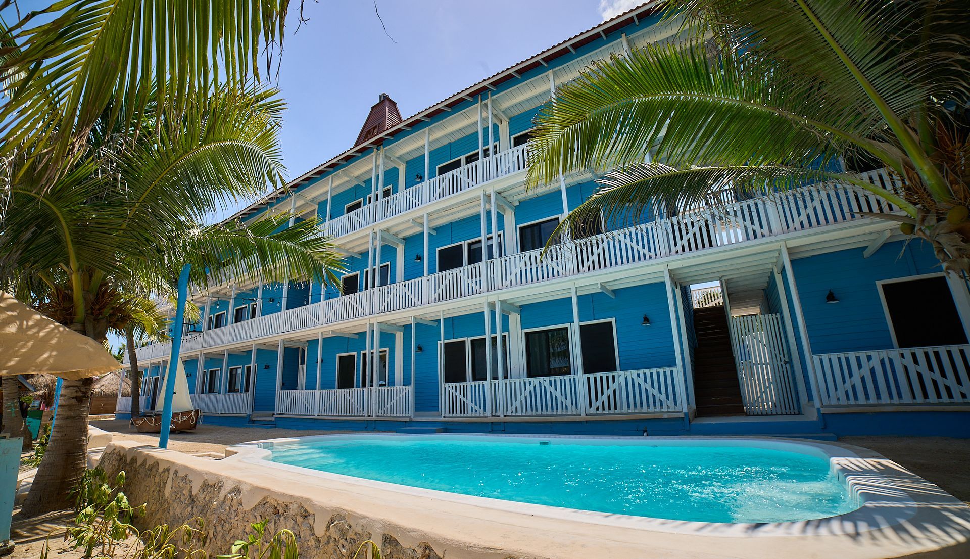 Hotel azul frente a la playa con balcones blancos y piscina, enmarcado por palmeras bajo un cielo soleado.