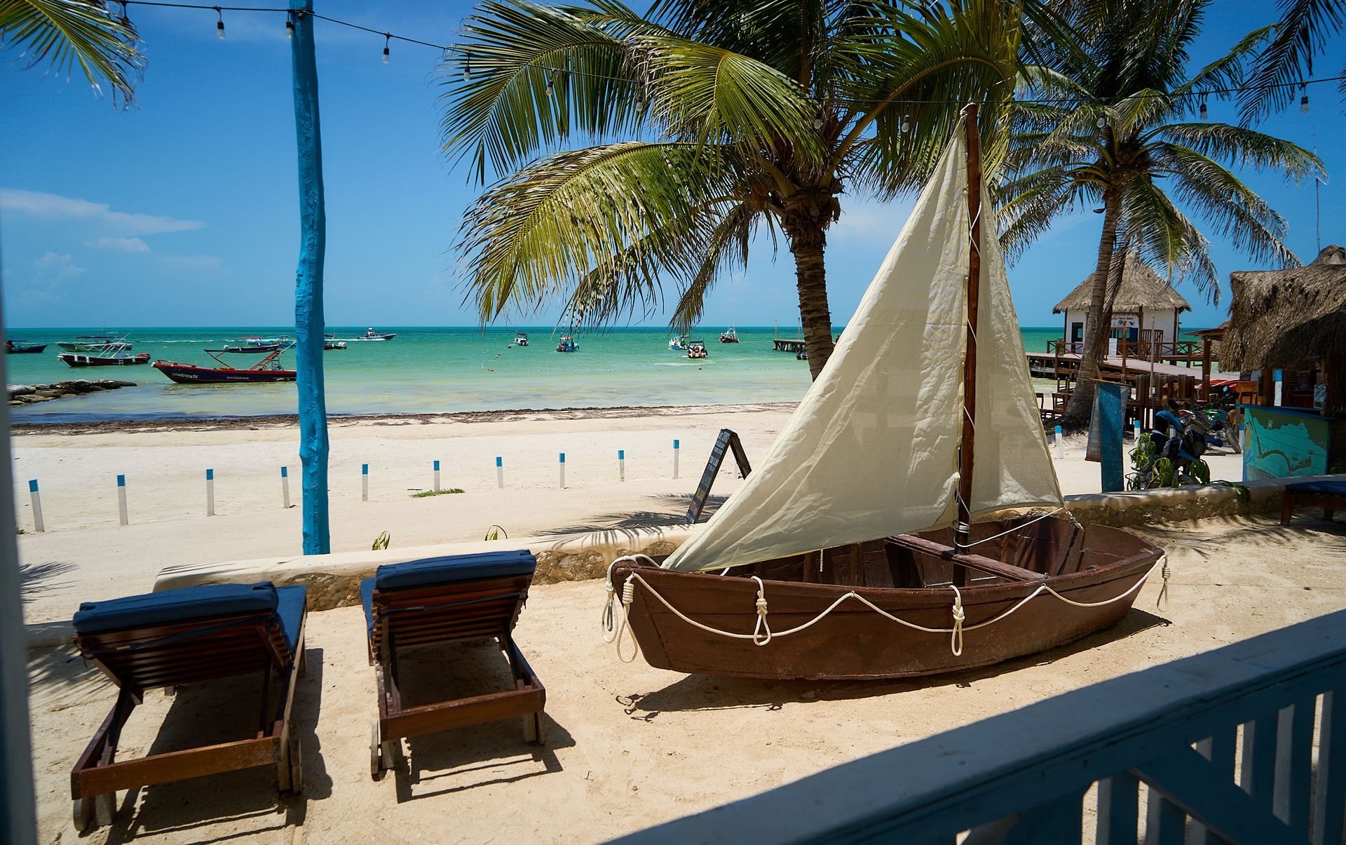 Escena de playa con tumbonas, palmeras y un velero en aguas turquesas.