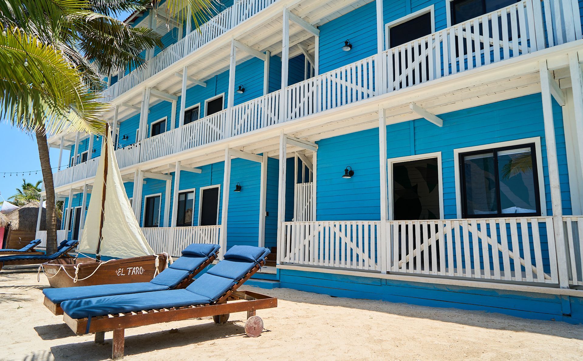 Edificio azul de un complejo turístico frente al mar, con balcones blancos y tumbonas en una playa de arena.