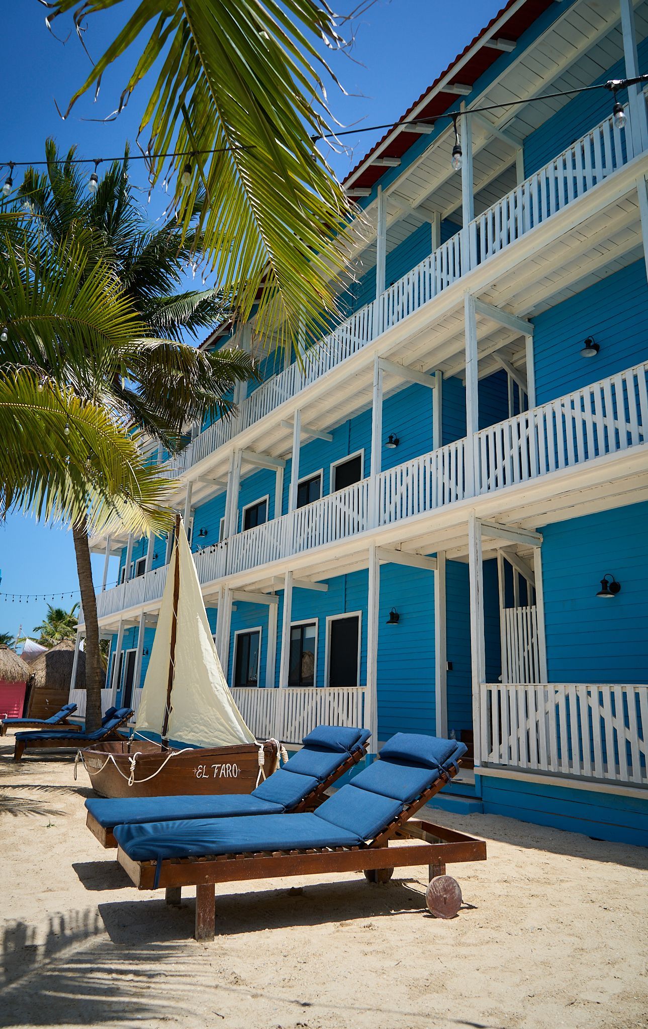 Hotel azul frente a la playa con balcones blancos y tumbonas en un patio de arena.