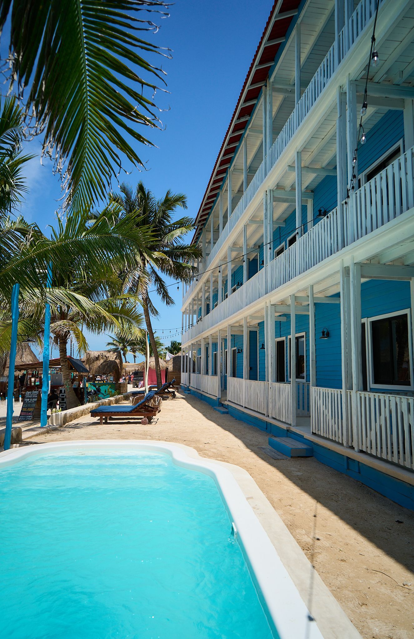 Piscina junto a un complejo turístico de playa de arena blanca y azul con palmeras y tumbonas bajo un cielo despejado.