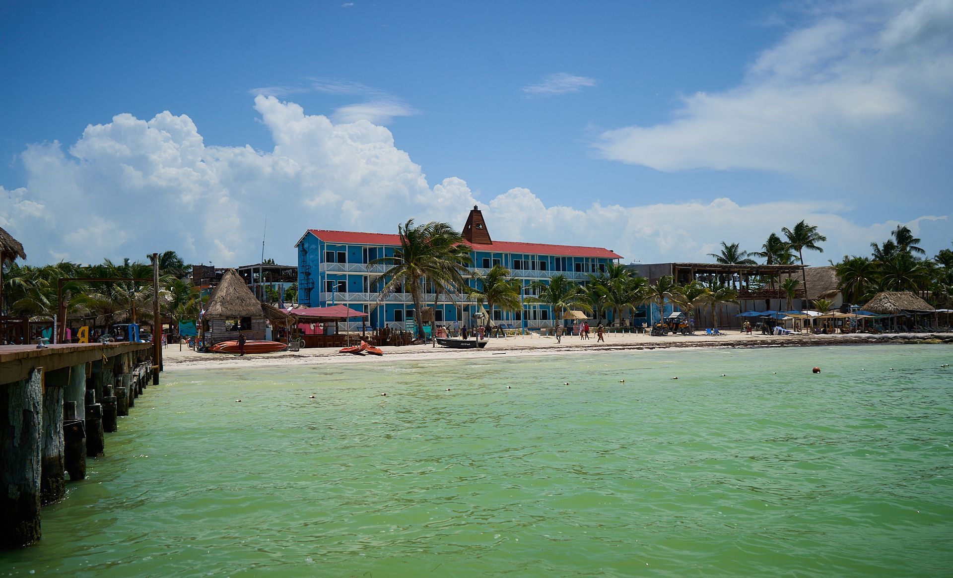 Complejo turístico tropical frente al mar con edificios azules y rojos, palmeras, playa de arena y aguas verdes tranquilas.
