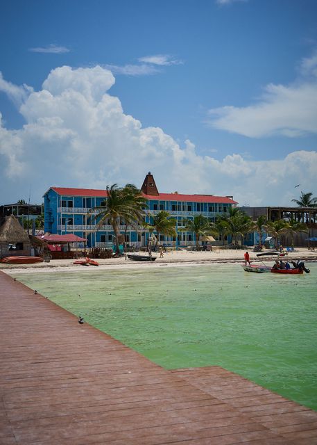 Complejo turístico frente al mar con edificio azul, playa de arena, agua verde y un muelle rojo bajo un cielo azul.
