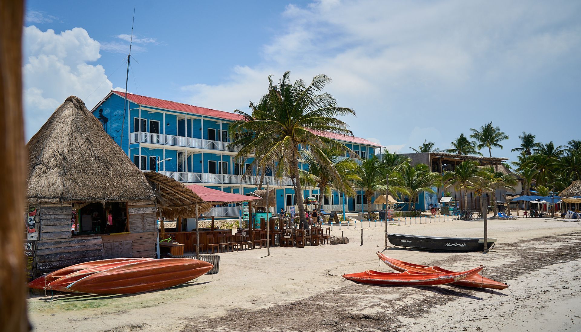 Complejo turístico frente a la playa con cabañas de paja, edificio azul, palmeras y kayaks en la orilla arenosa.