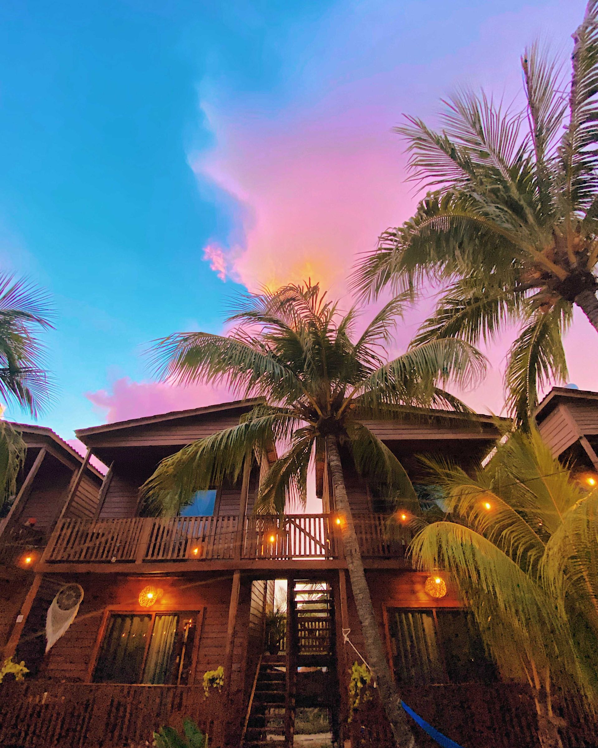 Casa tropical con palmeras al atardecer bajo un cielo rosa y azul, luces cálidas brillando en la fachada.