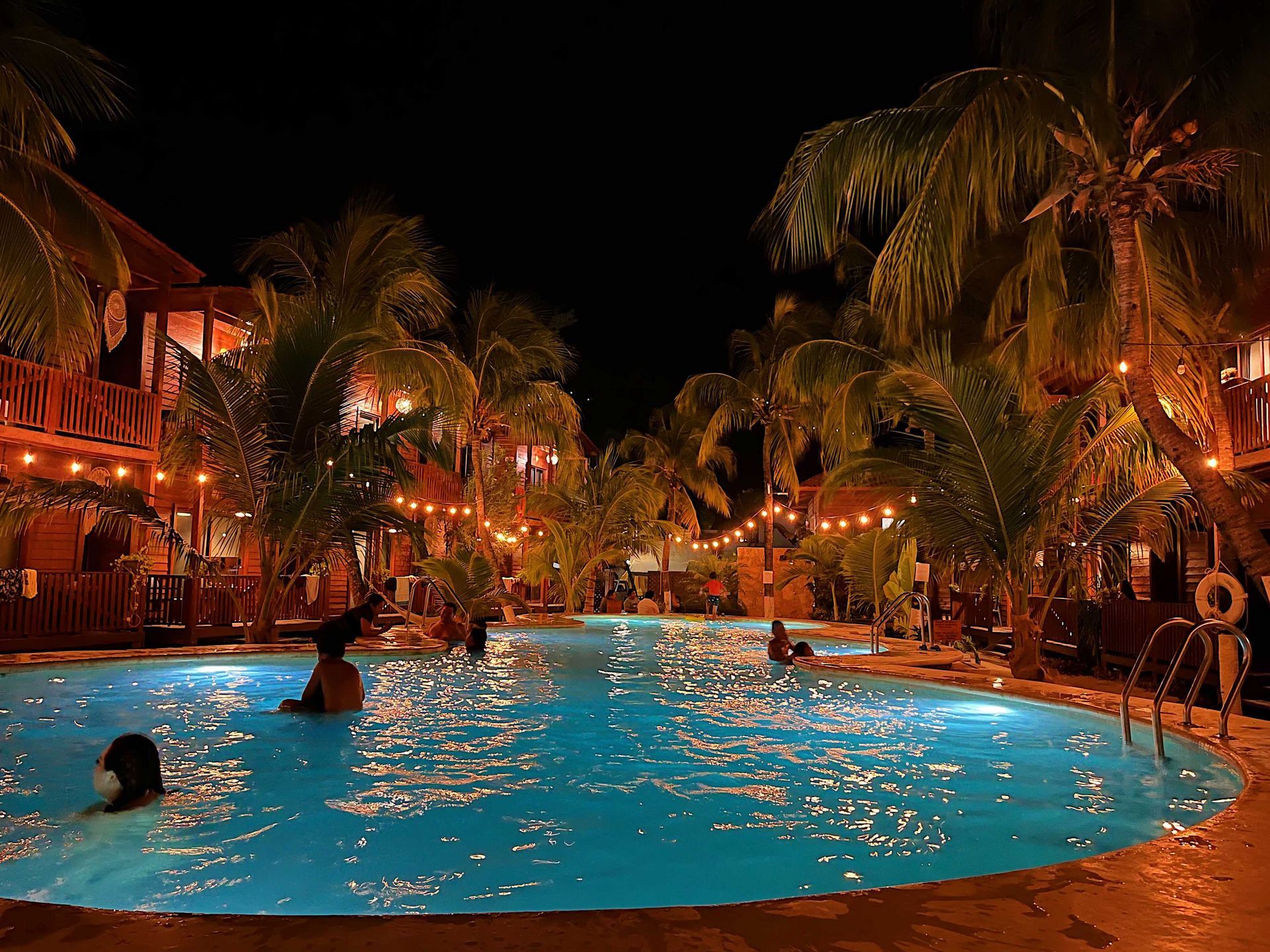 Piscina nocturna de un complejo turístico con bañistas, palmeras y luces naranjas cálidas que se reflejan en el agua azul.
