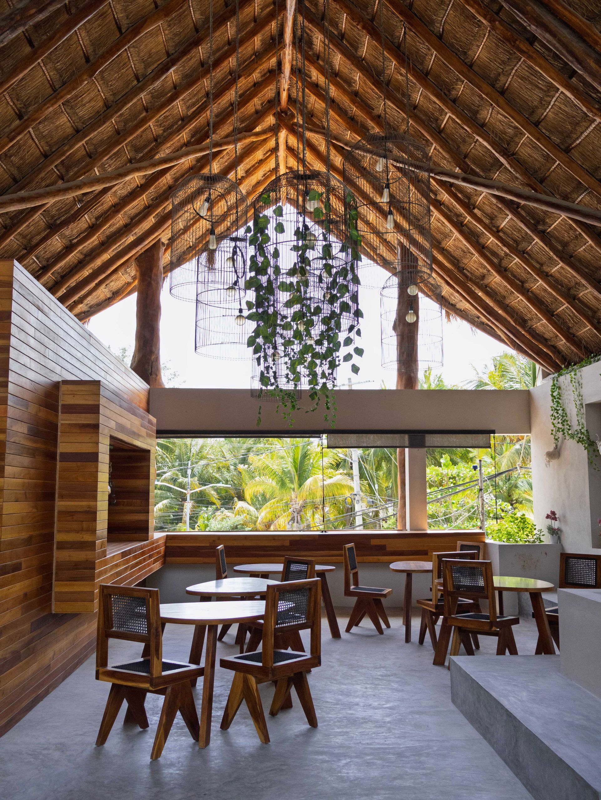 Cafetería al aire libre con mesas de madera bajo un alto techo de paja y exuberantes plantas verdes.