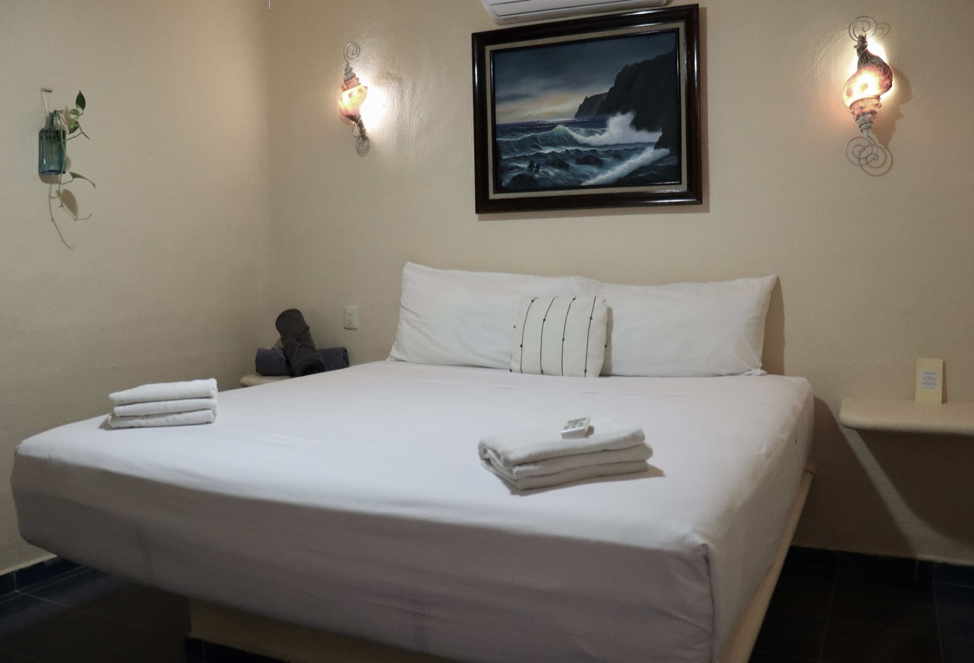 Cama bien hecha en una sencilla habitación de hotel con apliques de pared y cuadros con motivos marinos enmarcados.