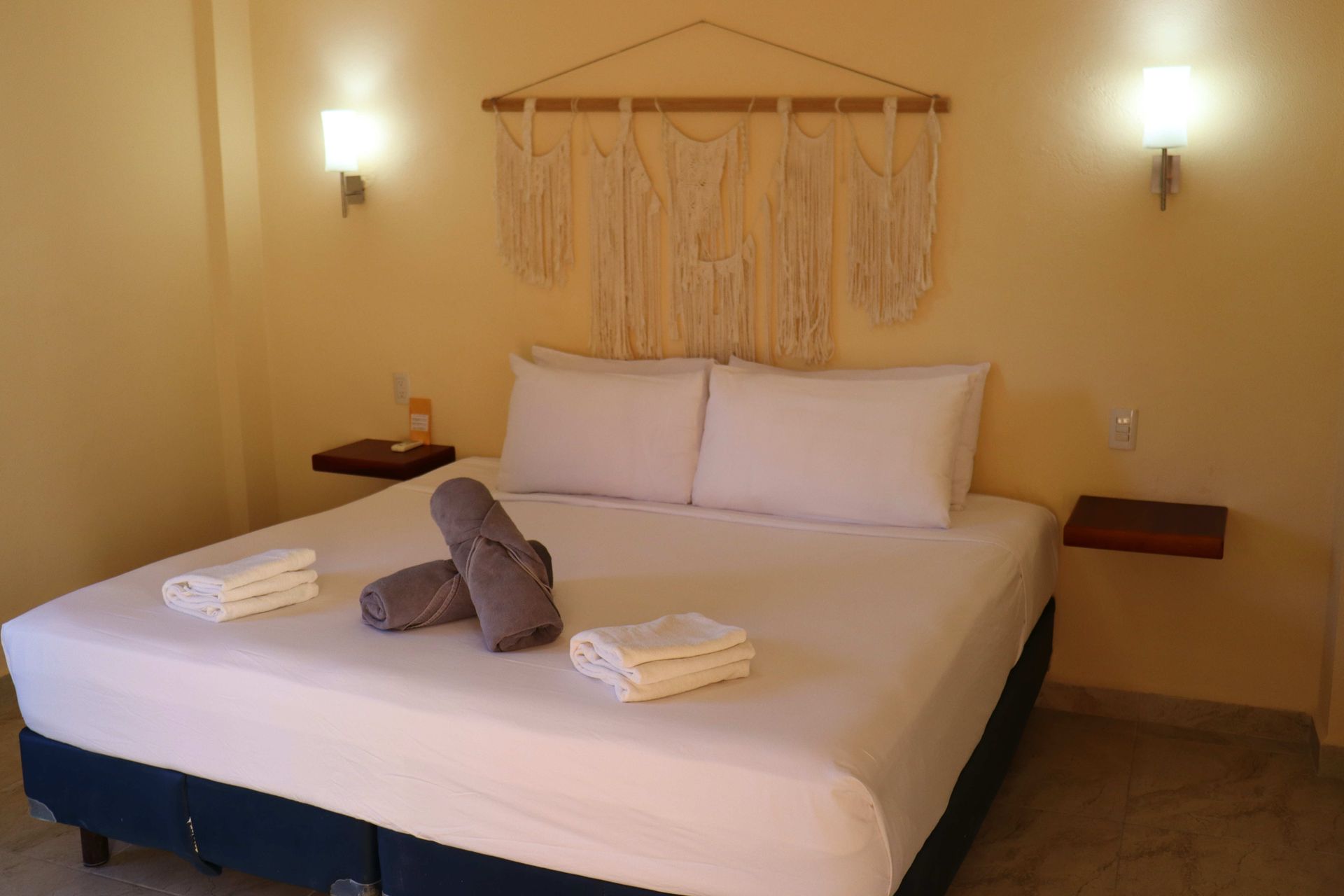 Cama bien hecha en un dormitorio de color beige con toallas dobladas y apliques de pared.