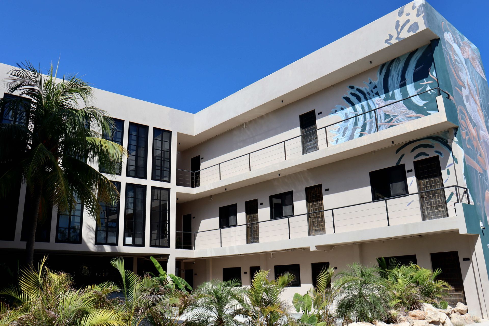 Edificio moderno de color blanco con detalles murales azules, balcones y exuberante vegetación bajo un cielo azul claro.