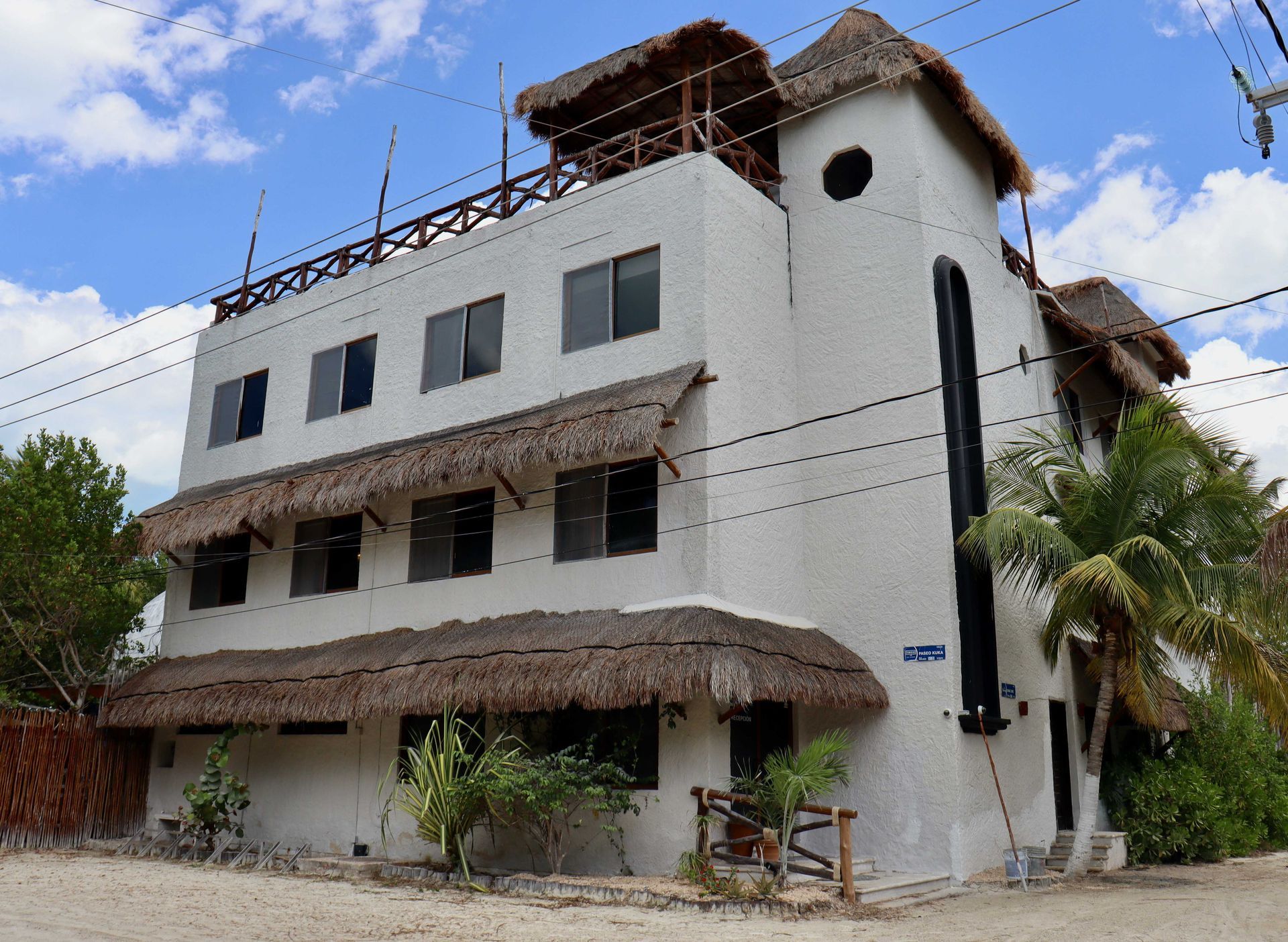 Edificio blanco de tres plantas con toldos de paja y estructura en la azotea bajo un cielo azul.