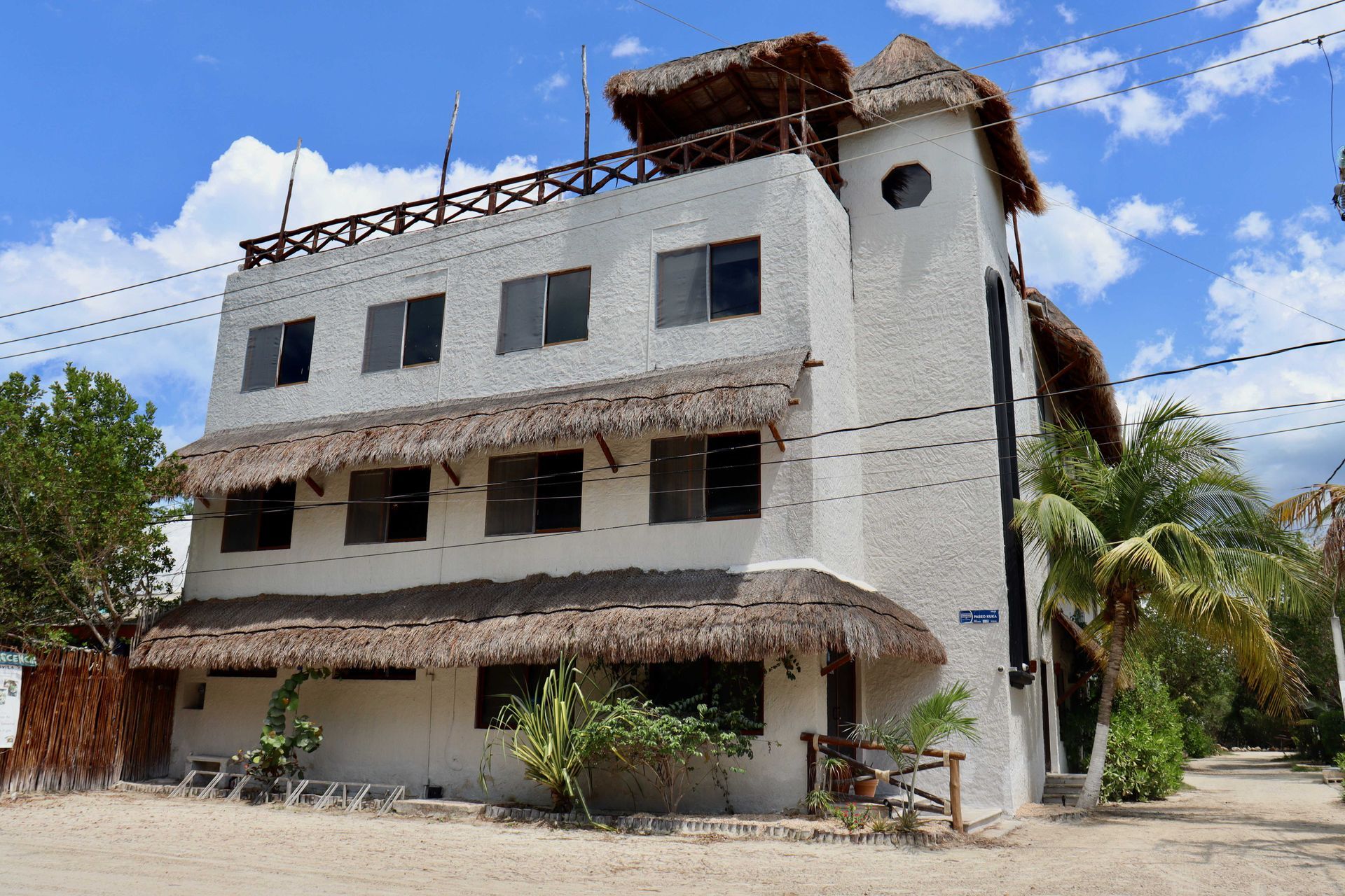 Edificio blanco de tres plantas, desgastado por el tiempo, con balcones y techo de paja bajo un cielo azul.