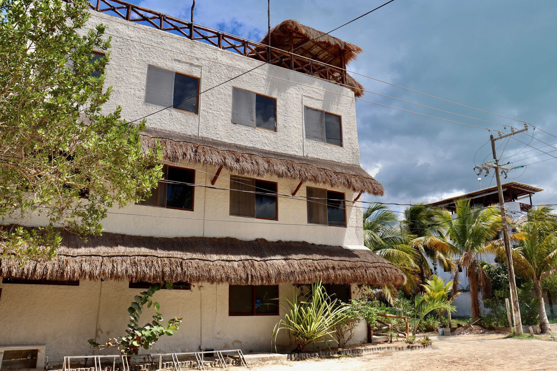 Edificio blanco de tres plantas con toldos de paja, palmeras y un cielo nublado.