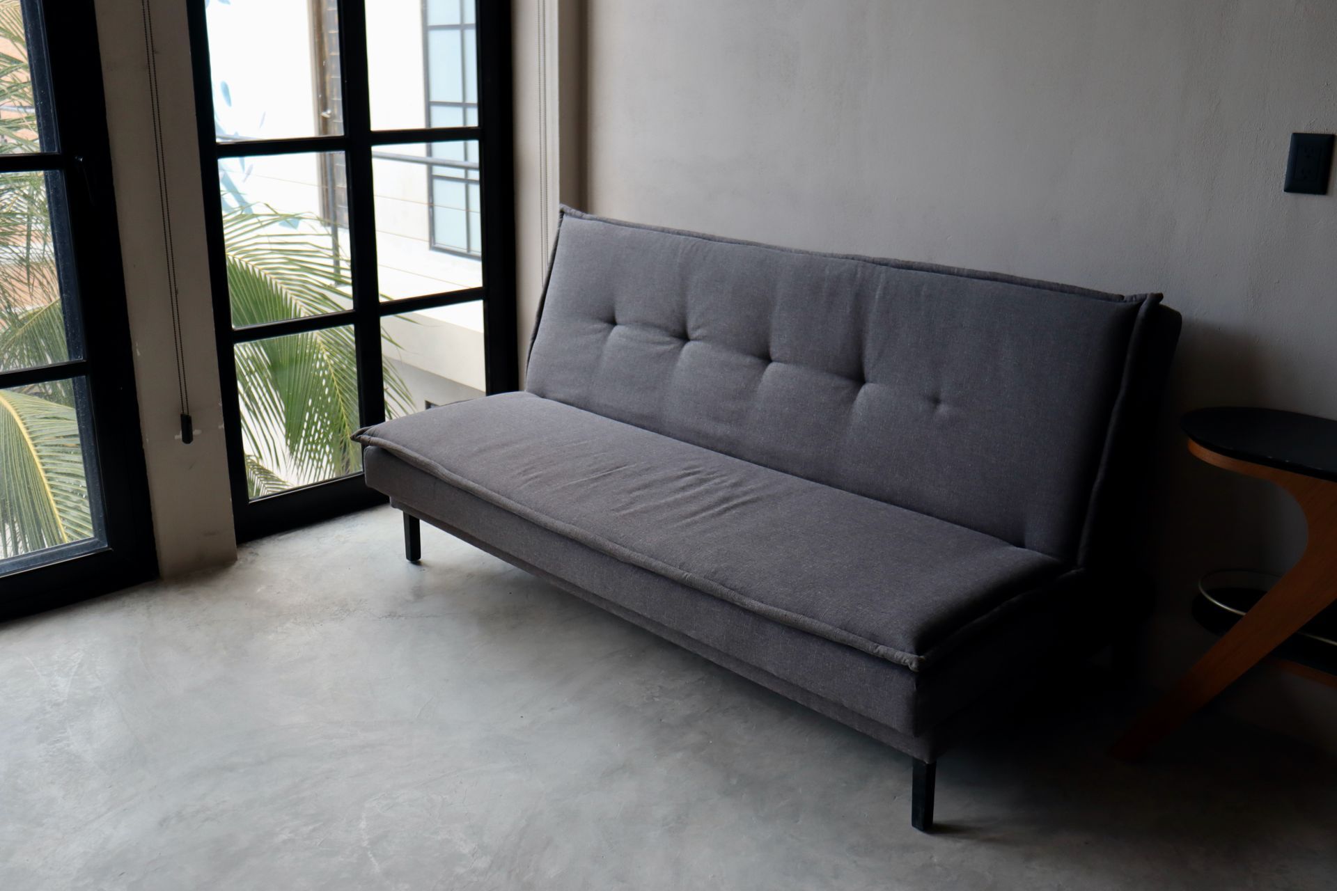 Sofá gris en una habitación minimalista junto a ventanas altas y una pequeña mesa auxiliar de madera.