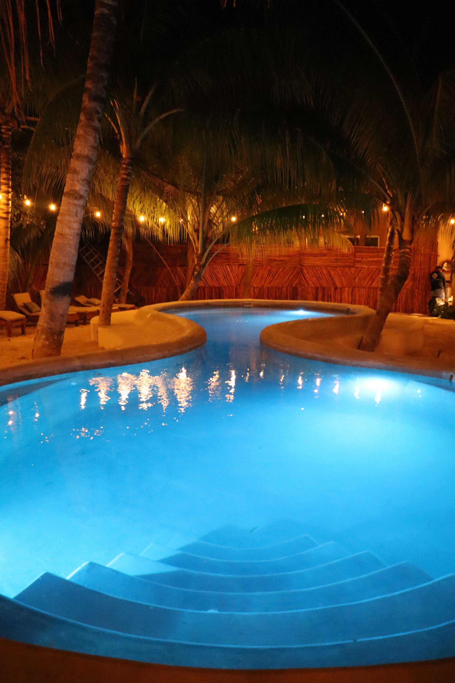 Piscina curva iluminada de azul por la noche, rodeada de palmeras y cálidas luces de resort.