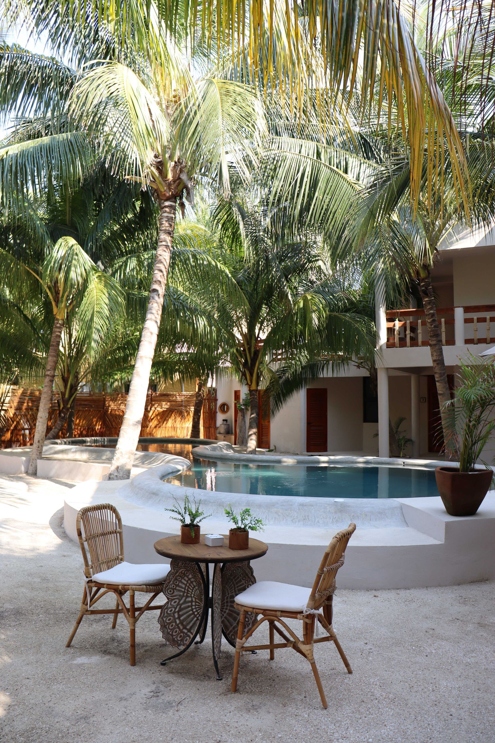 Piscina en el patio con palmeras, zona de descanso blanca y sillas de mimbre alrededor de una mesa pequeña.