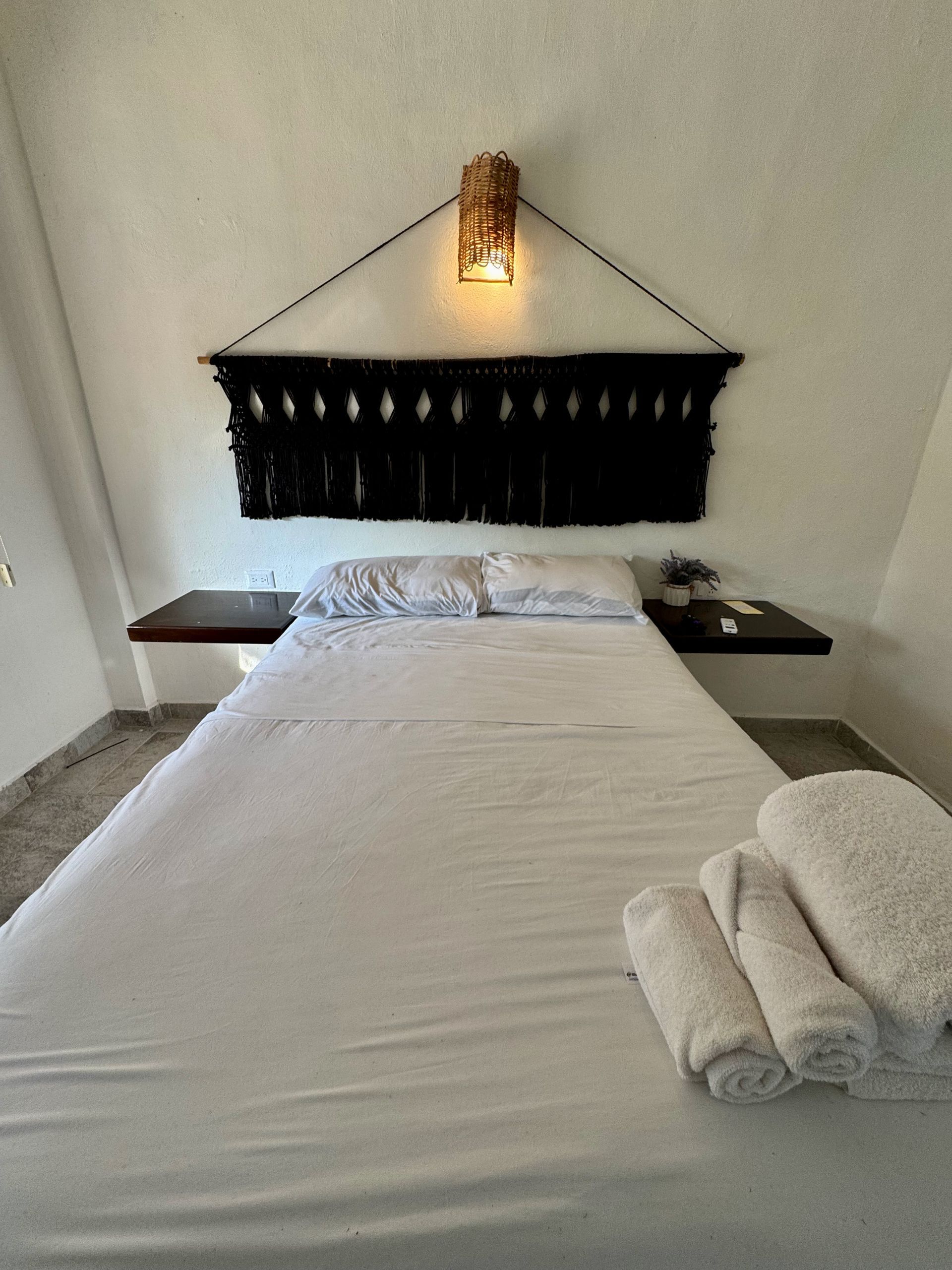 Habitación pequeña con una cama bien hecha, lámpara de pared, cabecero oscuro y toallas dobladas a los pies.