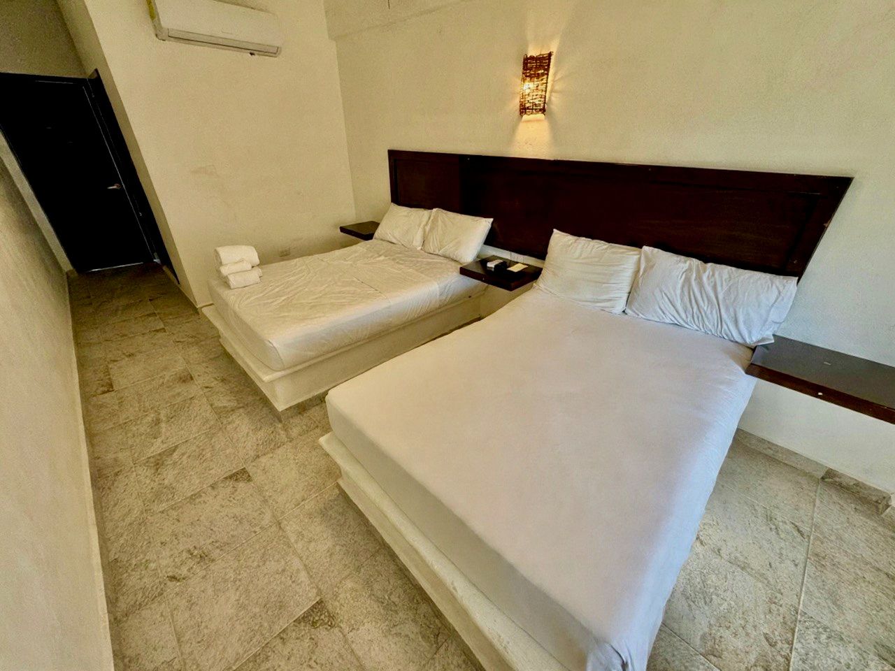 Habitación de hotel con dos camas blancas, cabecero de madera oscura y moqueta beige, vista desde la puerta.