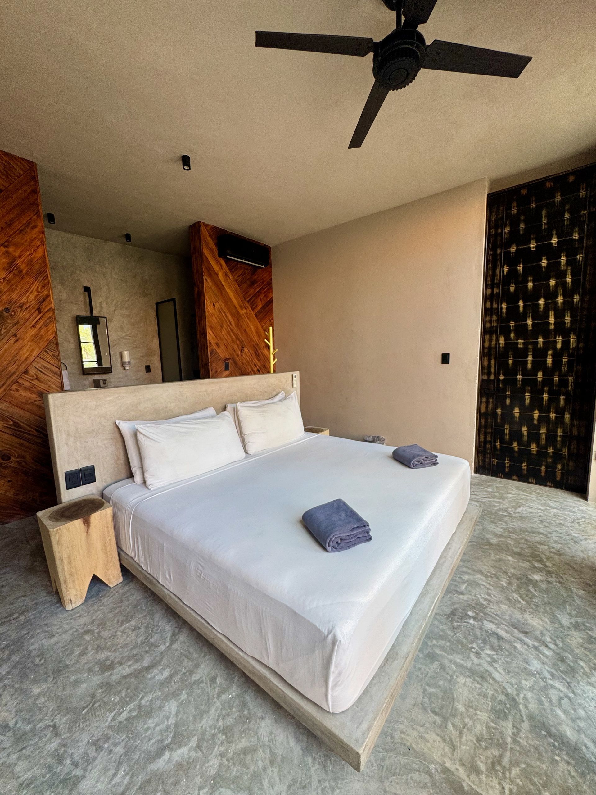 Dormitorio moderno con cama blanca, alfombra gris, ventilador de techo oscuro y paneles de pared estampados.
