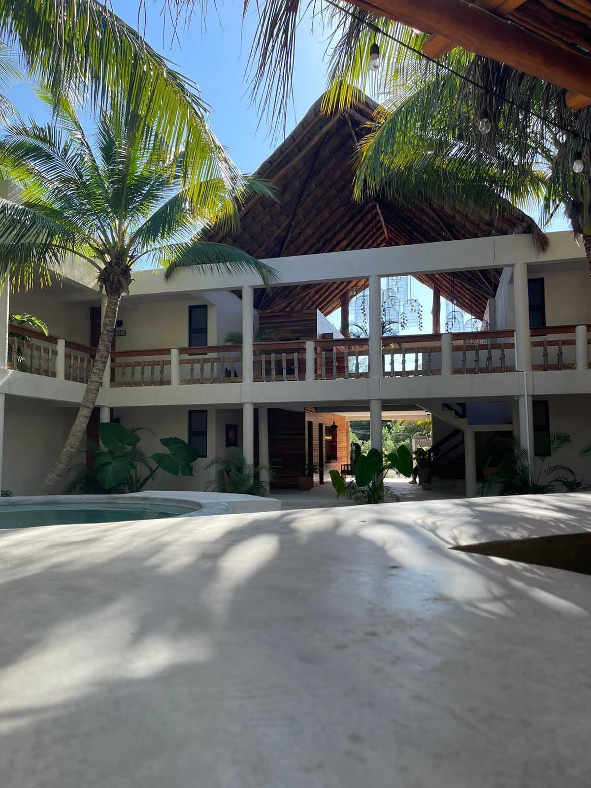 Patio de un complejo turístico tropical con un edificio blanco de dos plantas, palmeras y entrada con techo de paja.