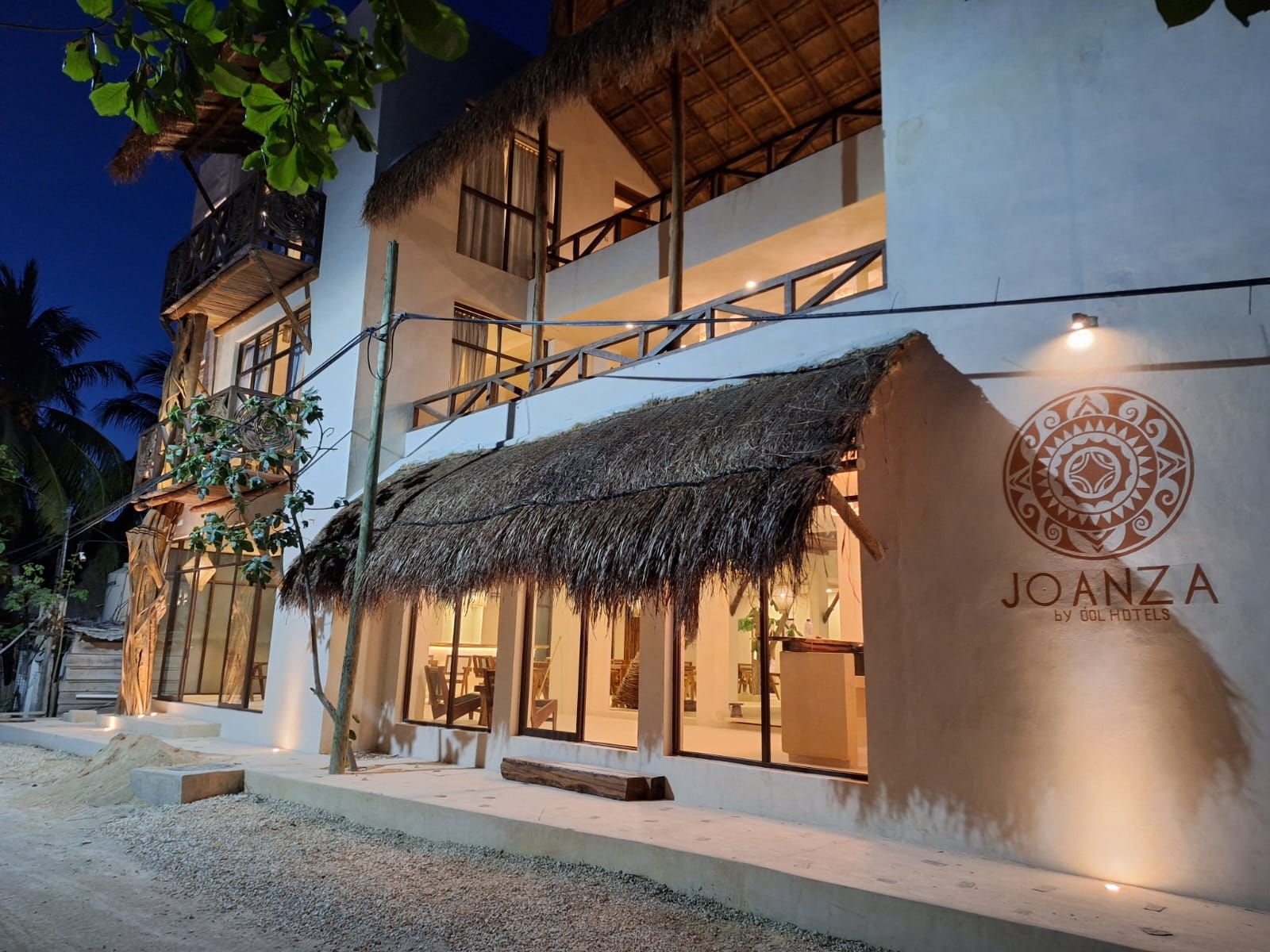 Exterior de un hotel frente a la playa con techo de paja al atardecer, con luces cálidas y un letrero luminoso.
