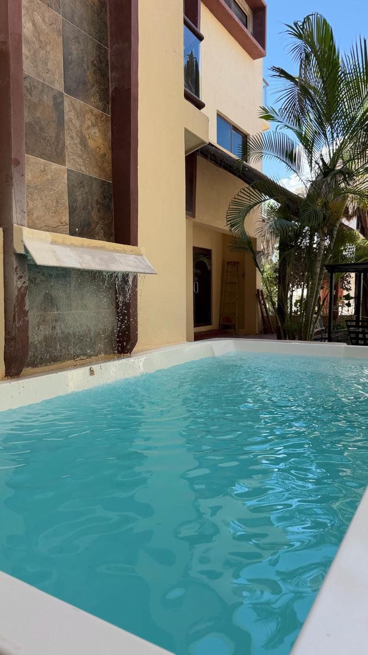 Piscina de color turquesa junto a un edificio de estuco y palmeras en un patio soleado.