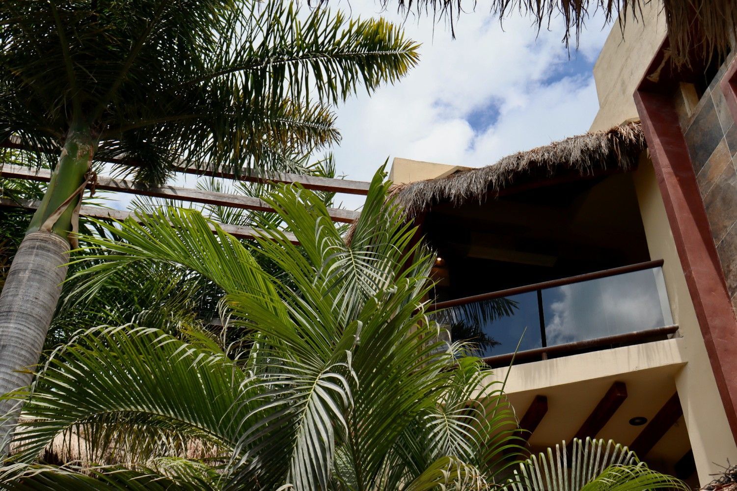 Edificio de un complejo turístico tropical con palmeras, techo de paja y balcones bajo un cielo parcialmente nublado.