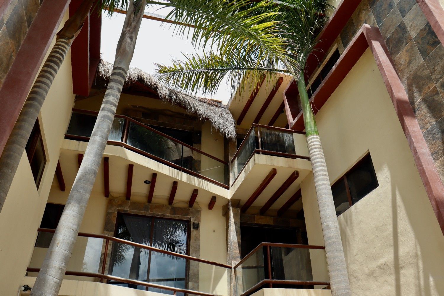 Edificio de varias plantas con paredes color crema, barandillas oscuras y palmeras visto desde abajo.