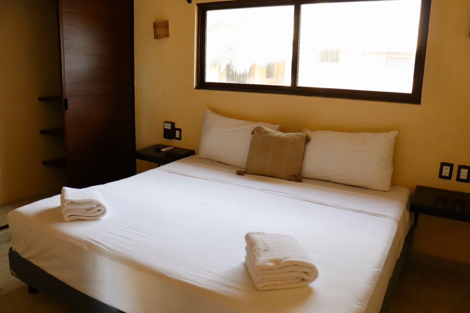 Habitación de hotel con una cama blanca bien hecha, paredes beige y una ventana sobre el cabecero.