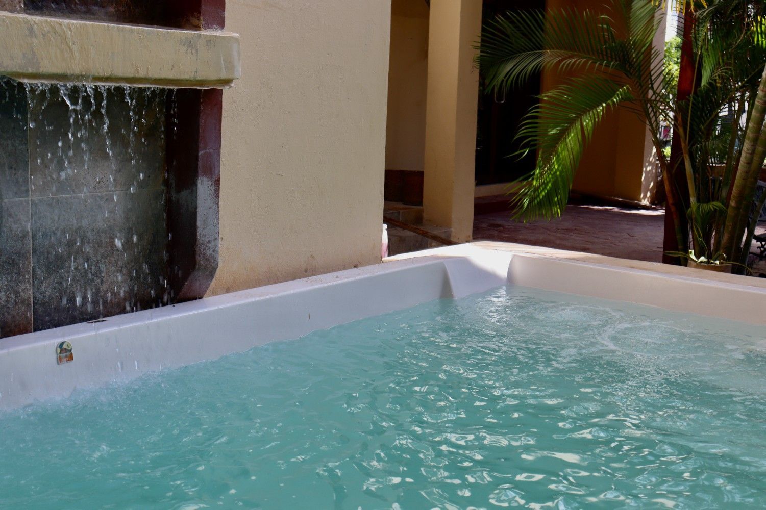 Pequeña piscina turquesa junto a un muro de patio beige con una fuente de agua burbujeante y palmeras.