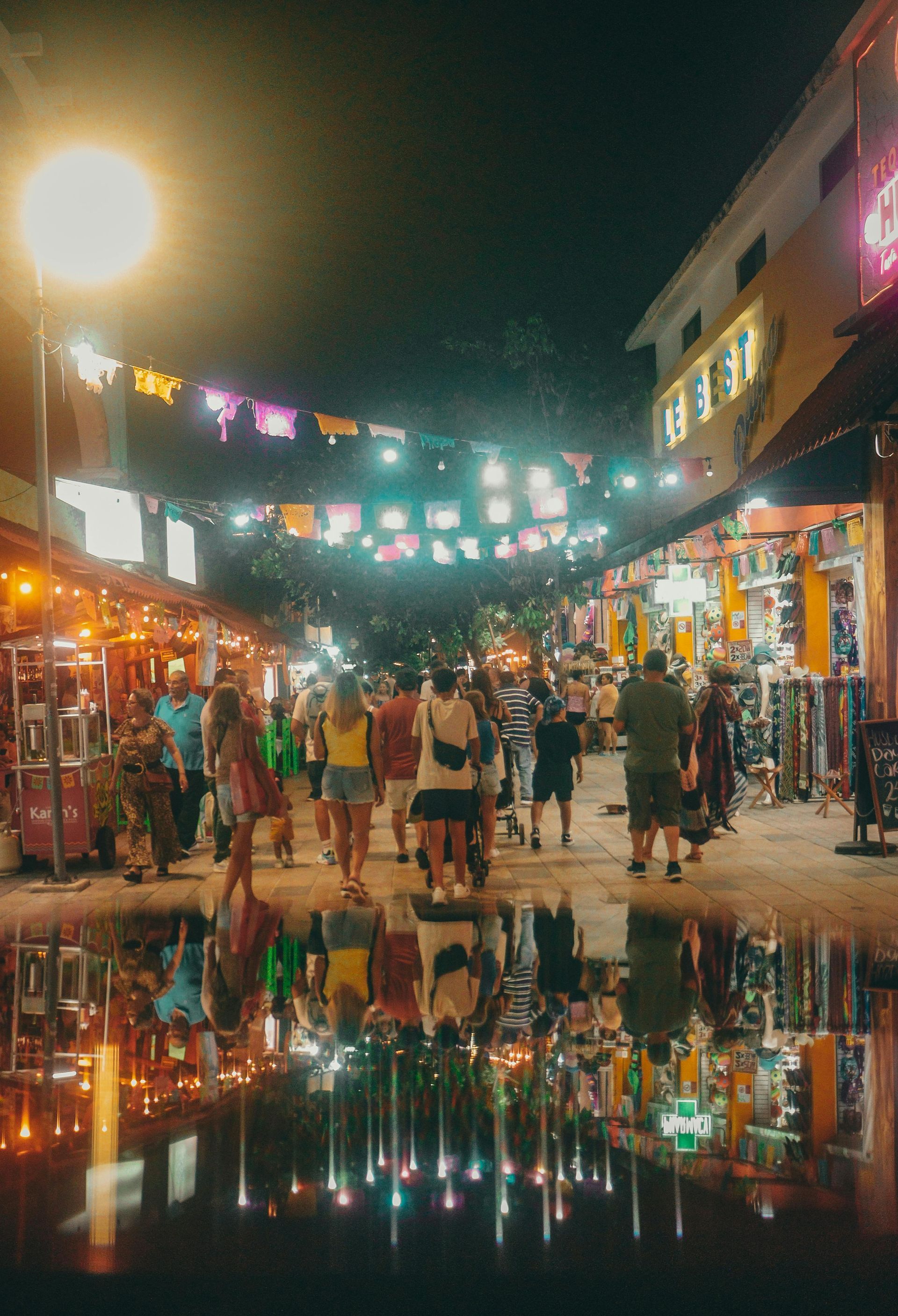 Calle del mercado nocturno con luces de colores, compradores atareados y reflejos brillantes en los charcos.