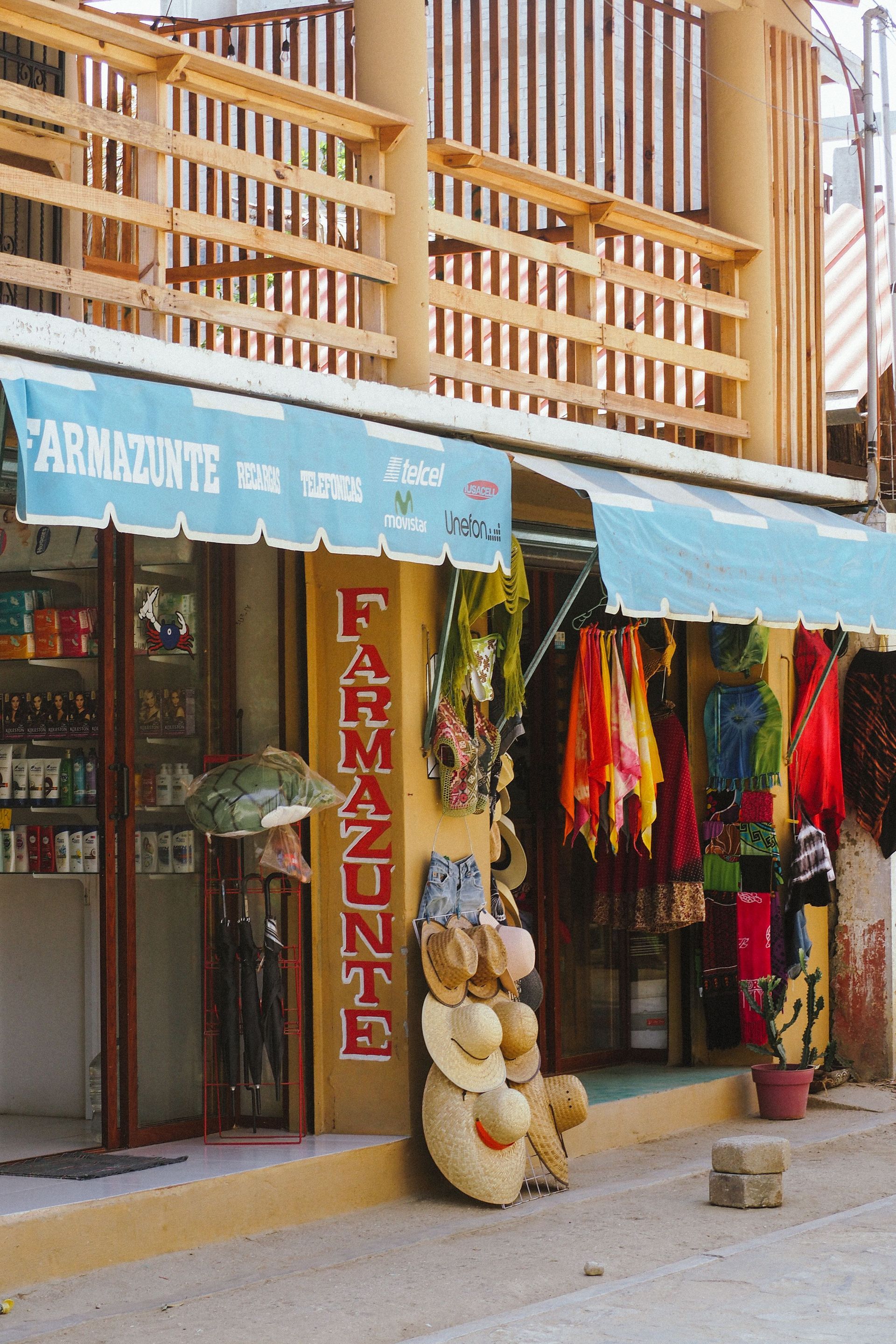 Fachada de una tienda con toldo azul, ropa colorida y una gran exhibición de ositos de peluche frente a una papelería.