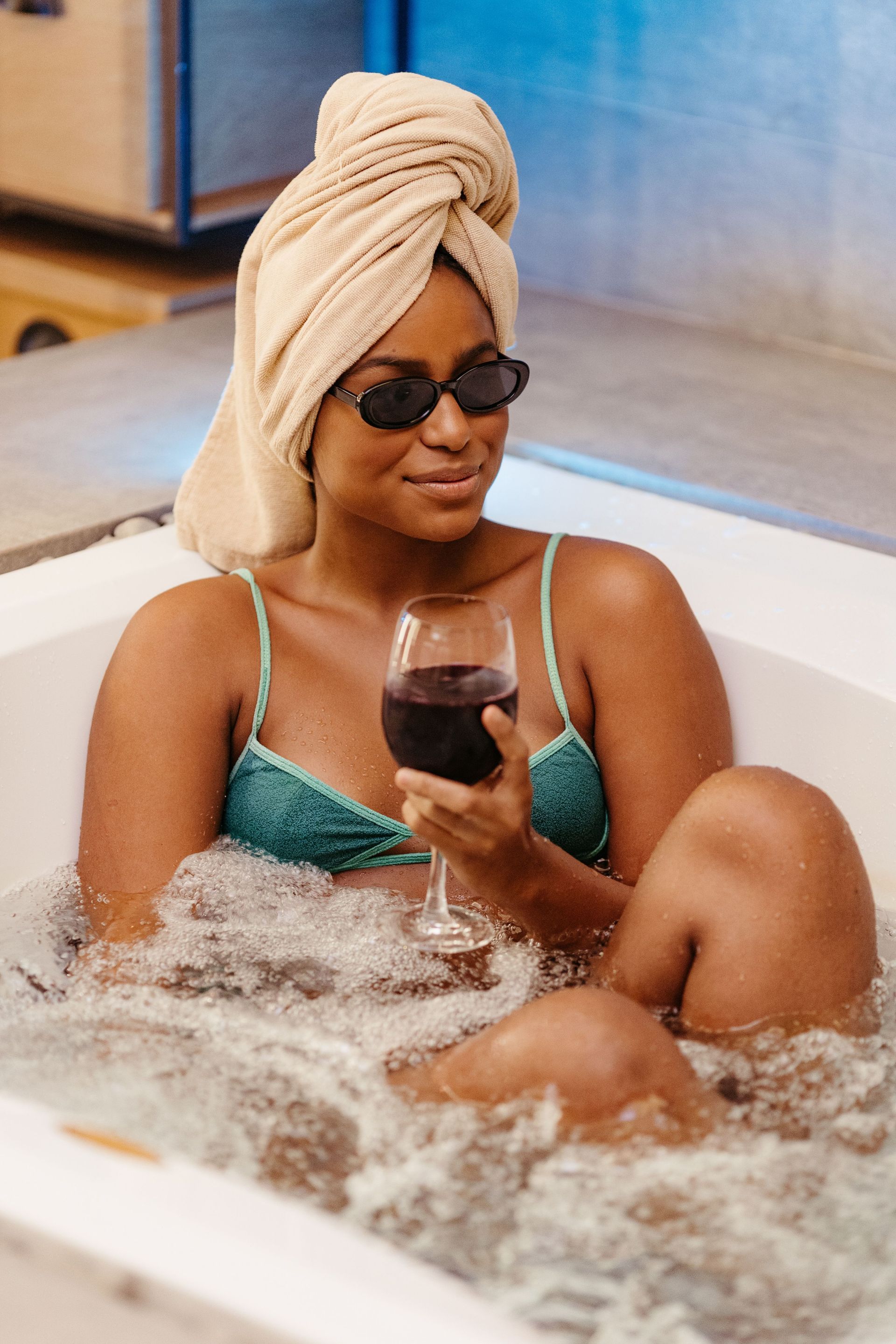 Una persona con un bañador turquesa y una toalla en la cabeza se relaja en un baño de burbujas, sosteniendo una copa de vino.