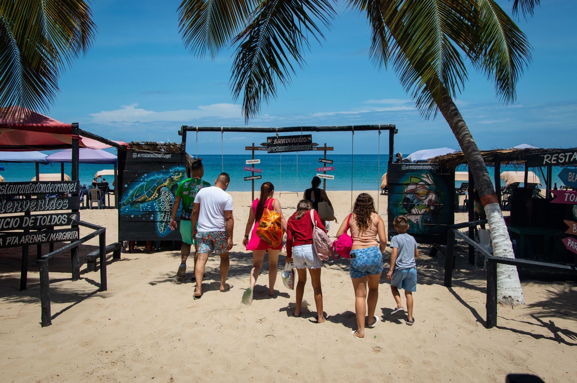 Gente caminando por la entrada de la playa hacia el océano, con palmeras y sombrillas sobre sus cabezas.