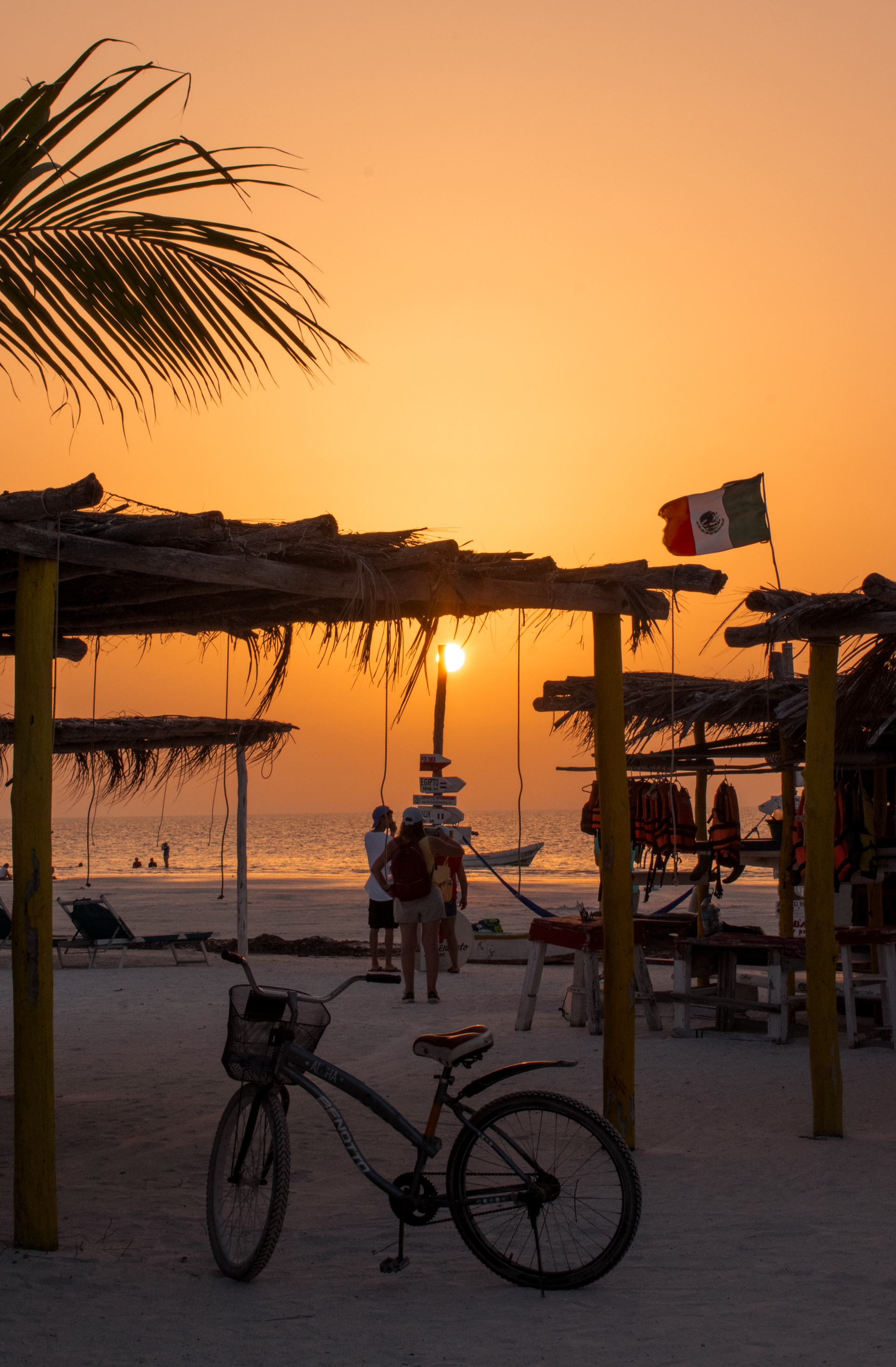 Escena de playa al atardecer con bicicletas, cabañas de palmeras y gente cerca de la orilla.