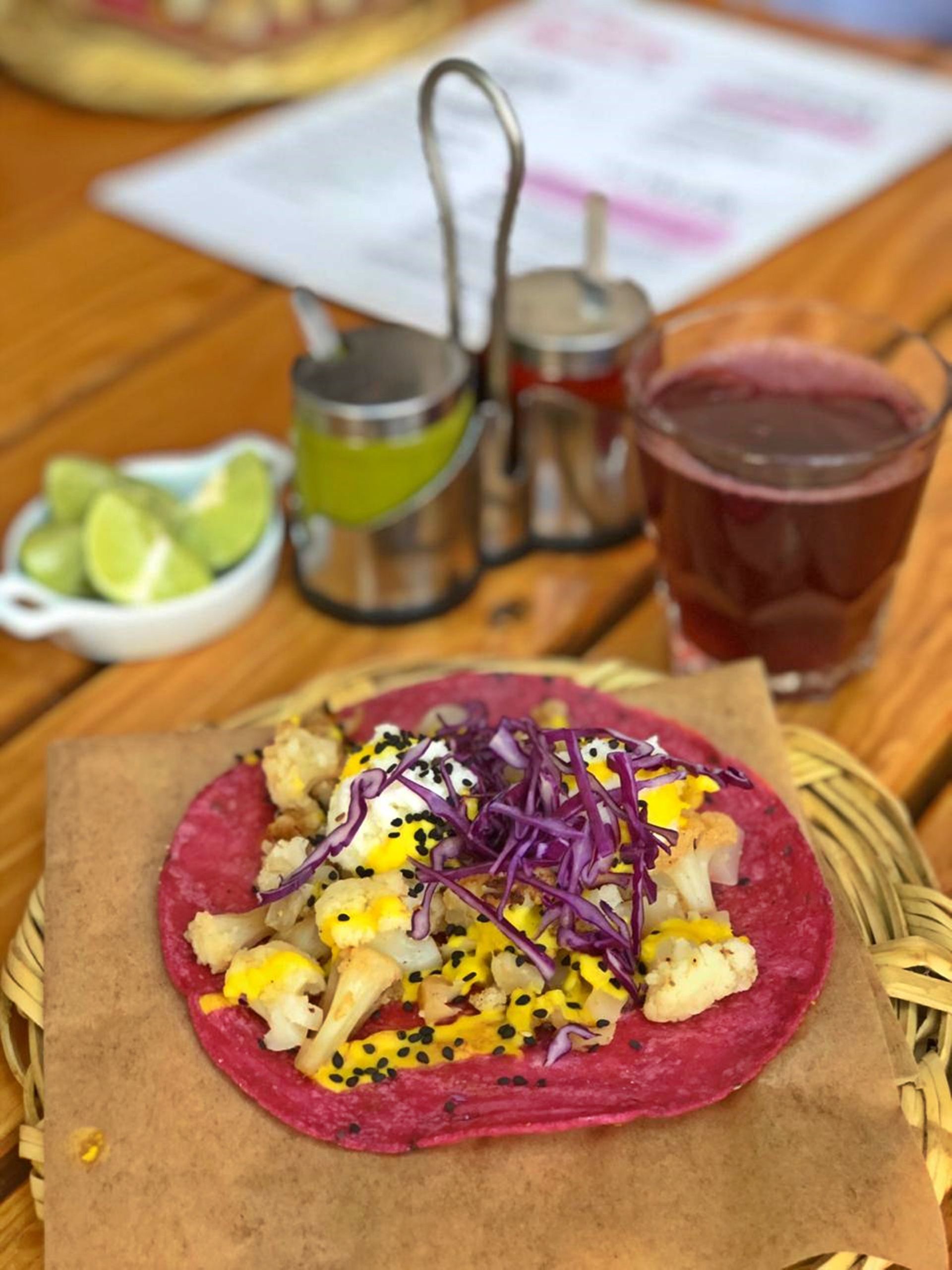 Taco colorido en una tortilla rosa con rodajas de lima y una bebida en una mesa de madera.
