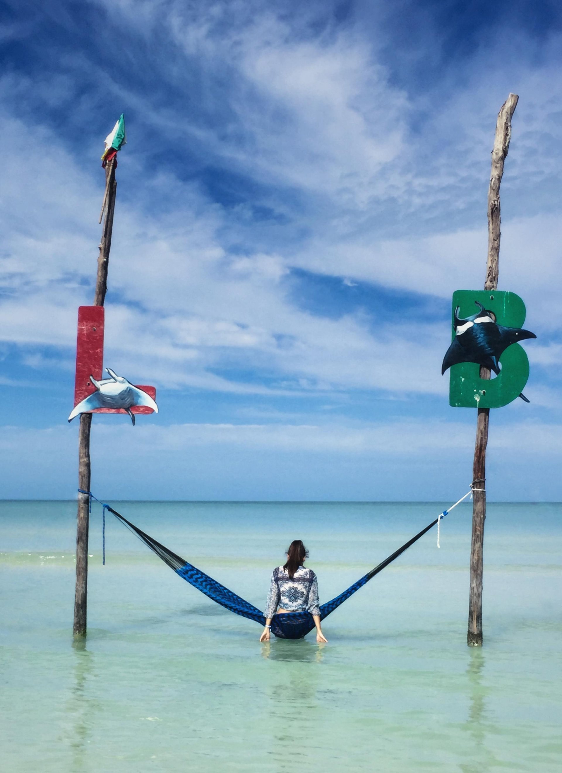 Una persona en una hamaca entre postes en aguas turquesas poco profundas bajo un cielo azul.
