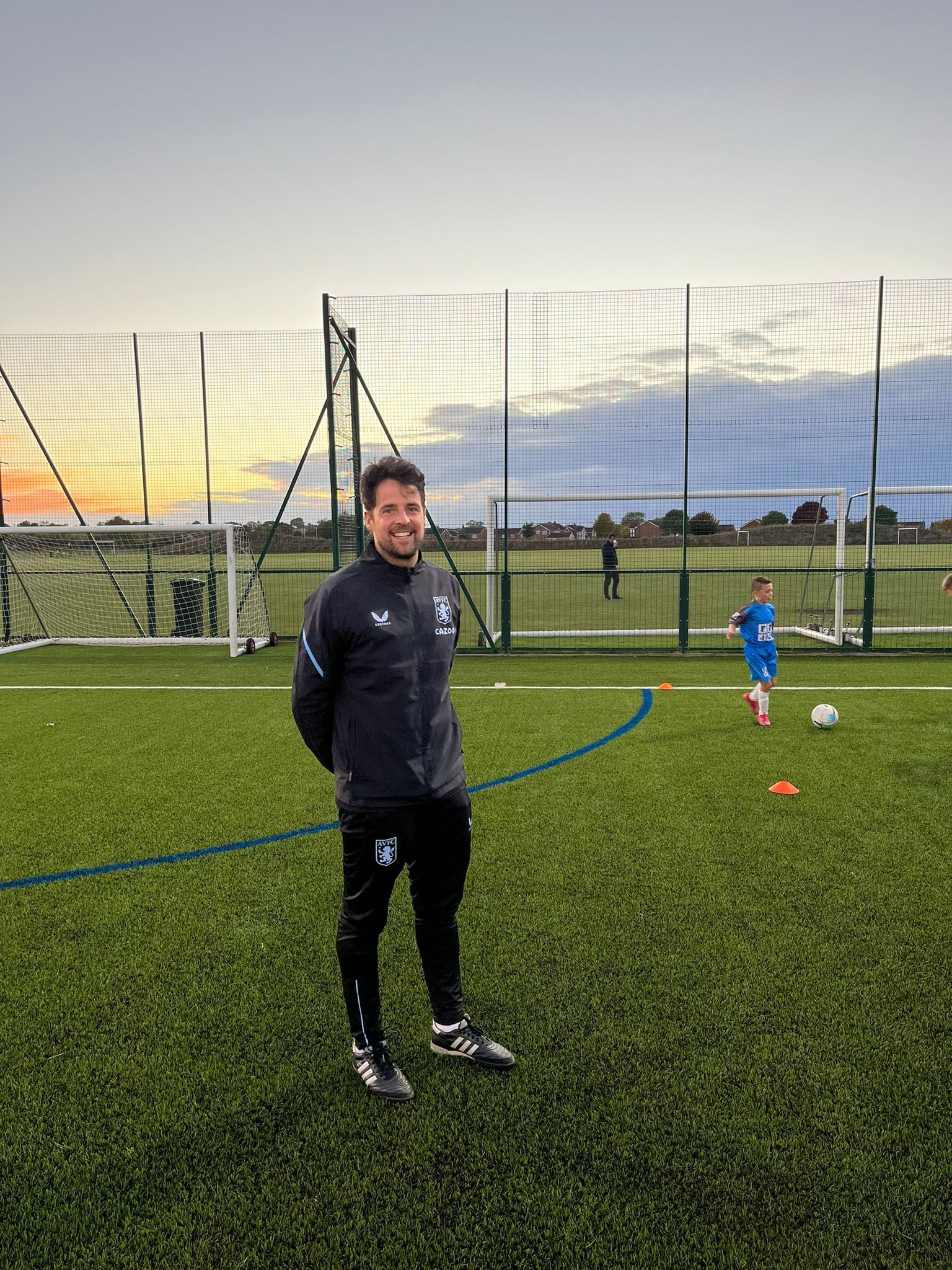 Coach in black sportswear standing on a football field at dusk, with a child and football in the background.