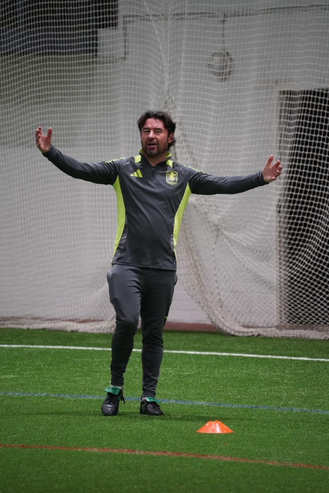Coach in gray tracksuit with arms outstretched on indoor turf beside an orange cone.
