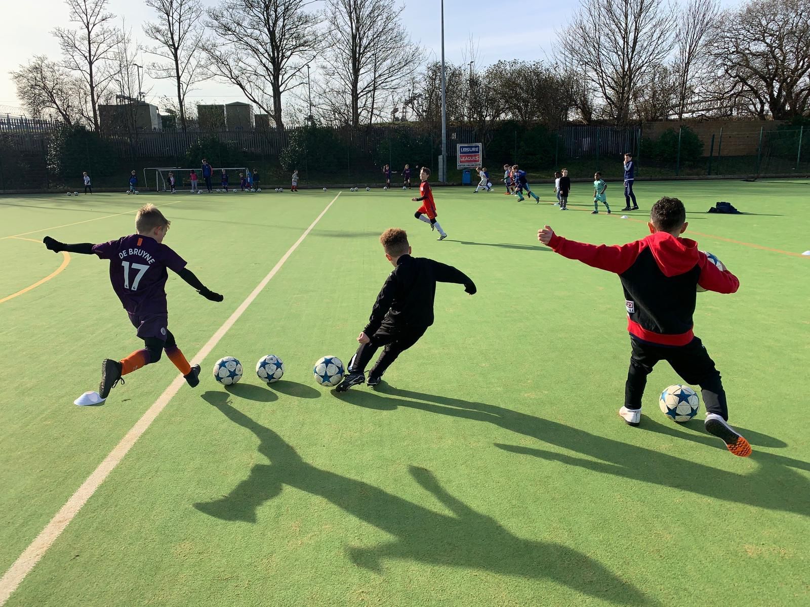 Children practicing football on a sunny field, with one kicking a ball toward a row of balls and cones.