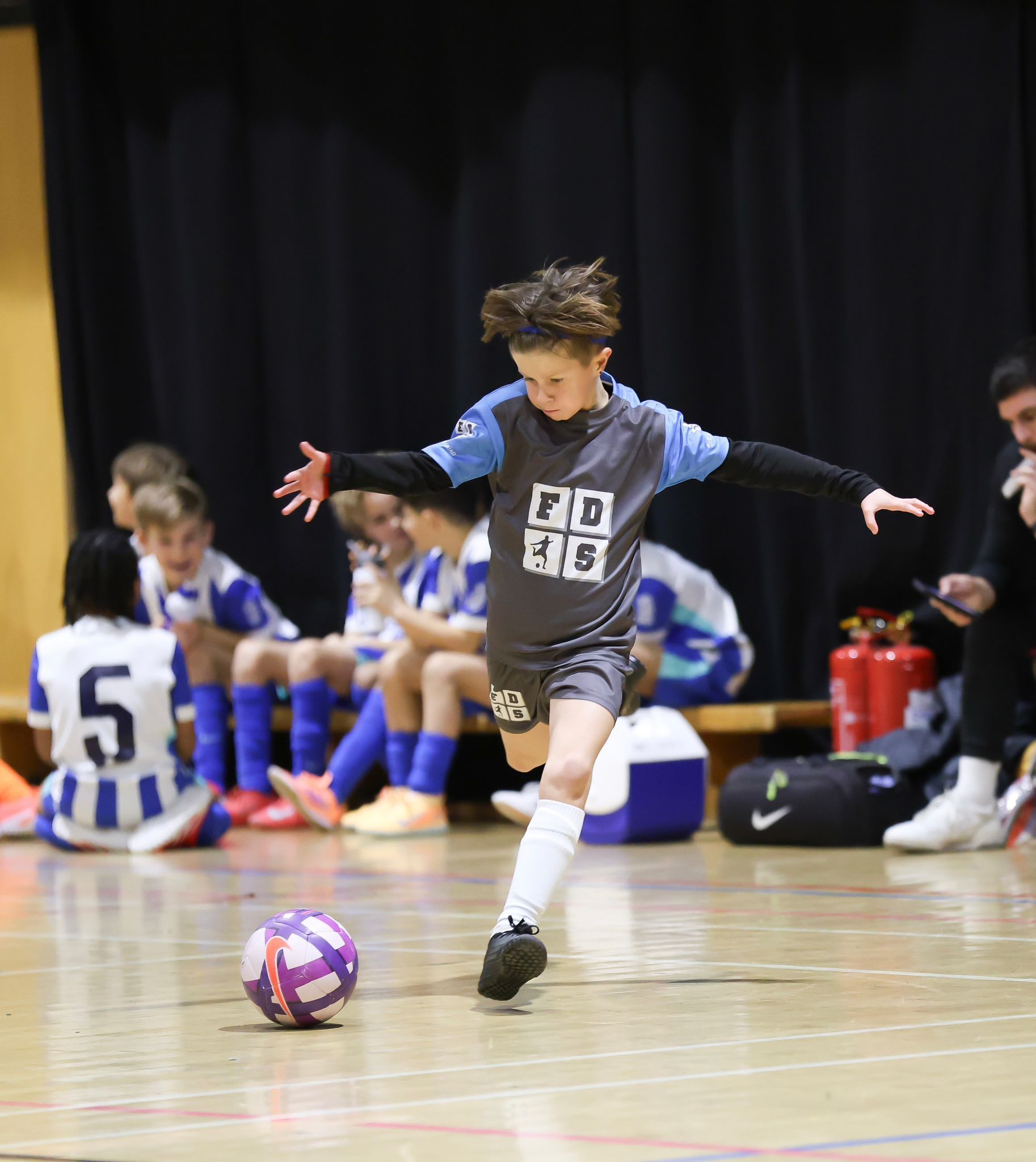 Young football player in blue dribbles a purple ball on an indoor court, teammates seated in the background