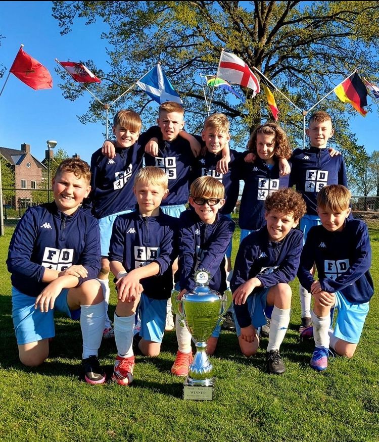 Youth football team posing on a sunny field with a trophy, wearing navy uniforms and holding flags of different countries behind them