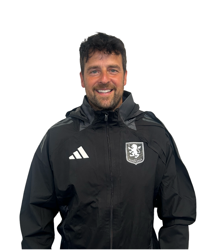 Coach in black sportswear standing on a football field at dusk, with a child and football in the background.